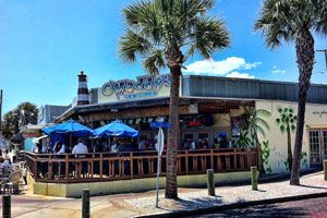 Daytime exterior of O'Maddy's — blue umbrellas, the turquoise wave sign, and palm trees on the Gulfport waterfront